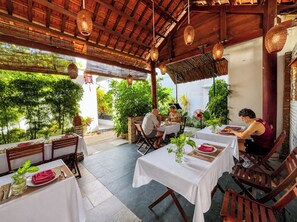 An outdoor dining area with tables and chairs, surrounded by greenery and wooden beams. - Trendy Life Villa Hoi An (Da Nang)
