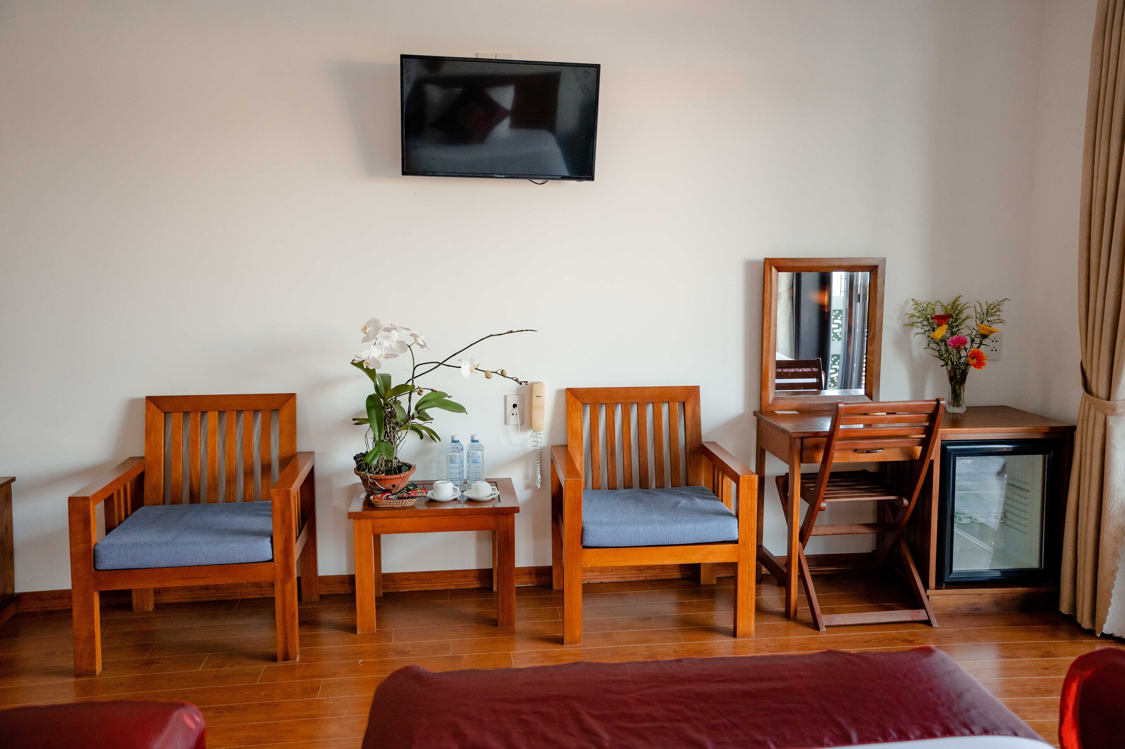 Habitación de hotel con dos sillas de madera, una mesita con una planta en maceta, un escritorio con un espejo, un televisor montado en la pared y un refrigerador pequeño.