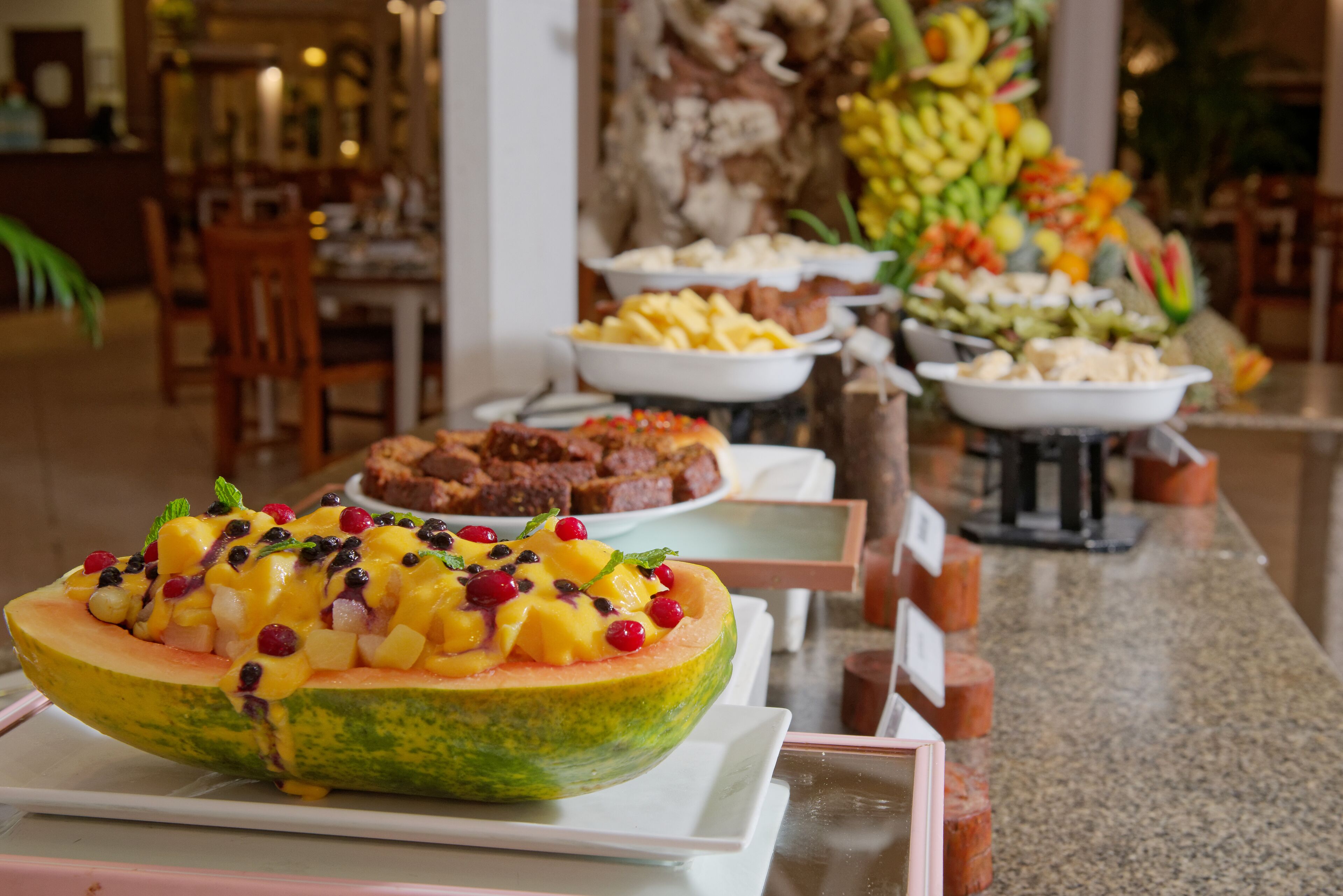 A buffet with various dishes, including a large melon bowl topped with fruit and berries, and other food items in the background.
