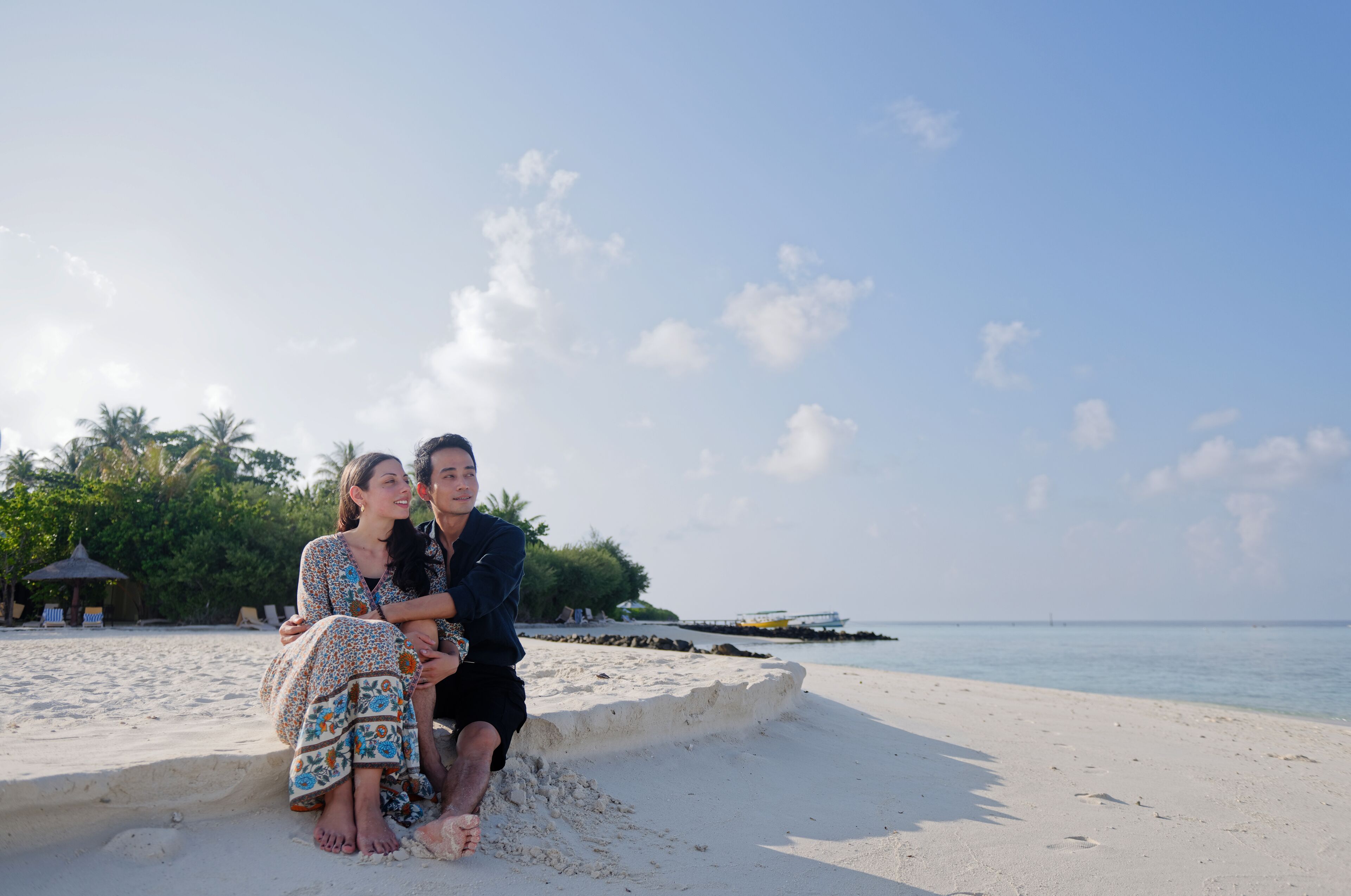 Una pareja sentada en una playa arenosa con cielo despejado y palmeras al fondo.