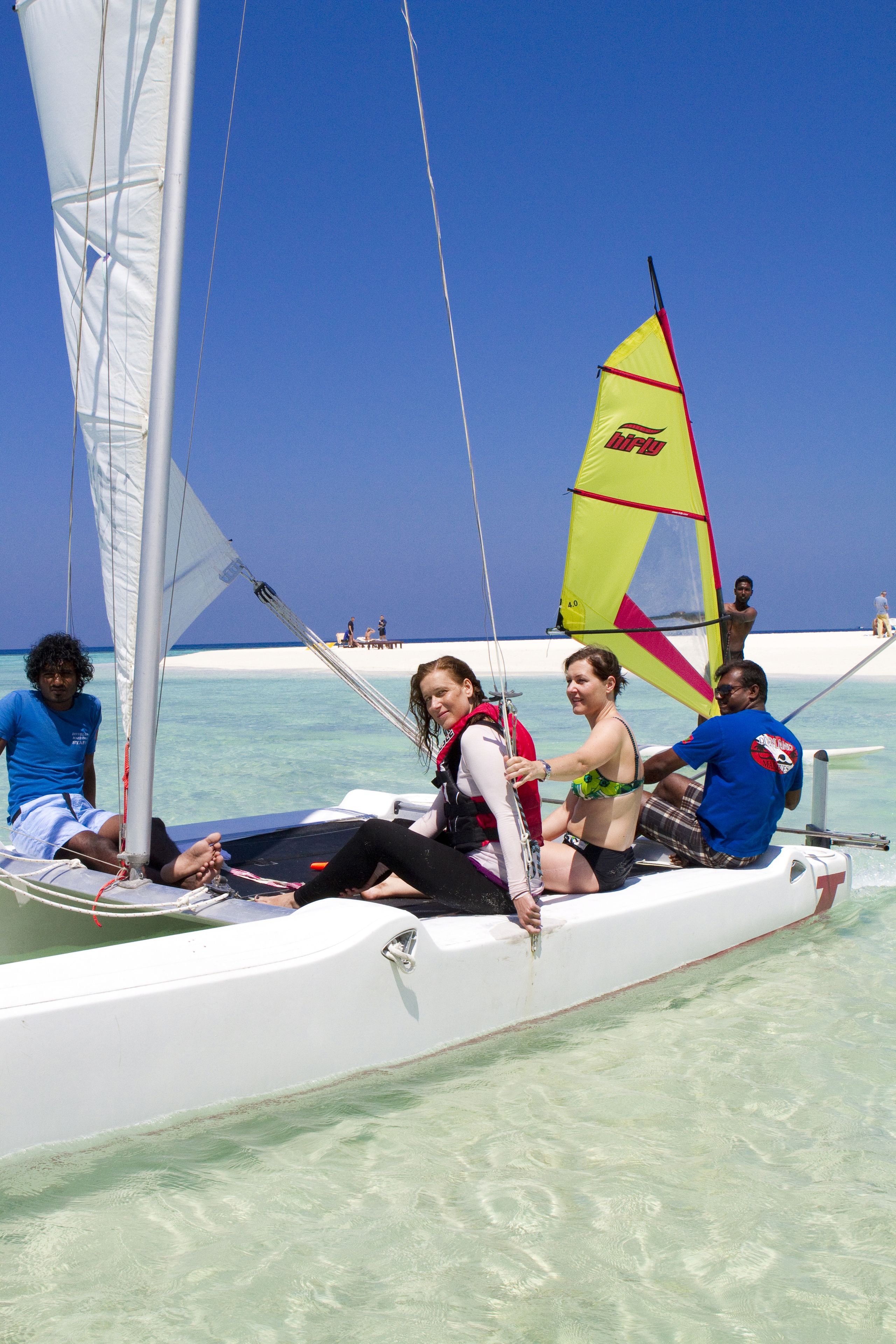Un velero con pasajeros, agua turquesa cristalina y una playa arenosa al fondo.