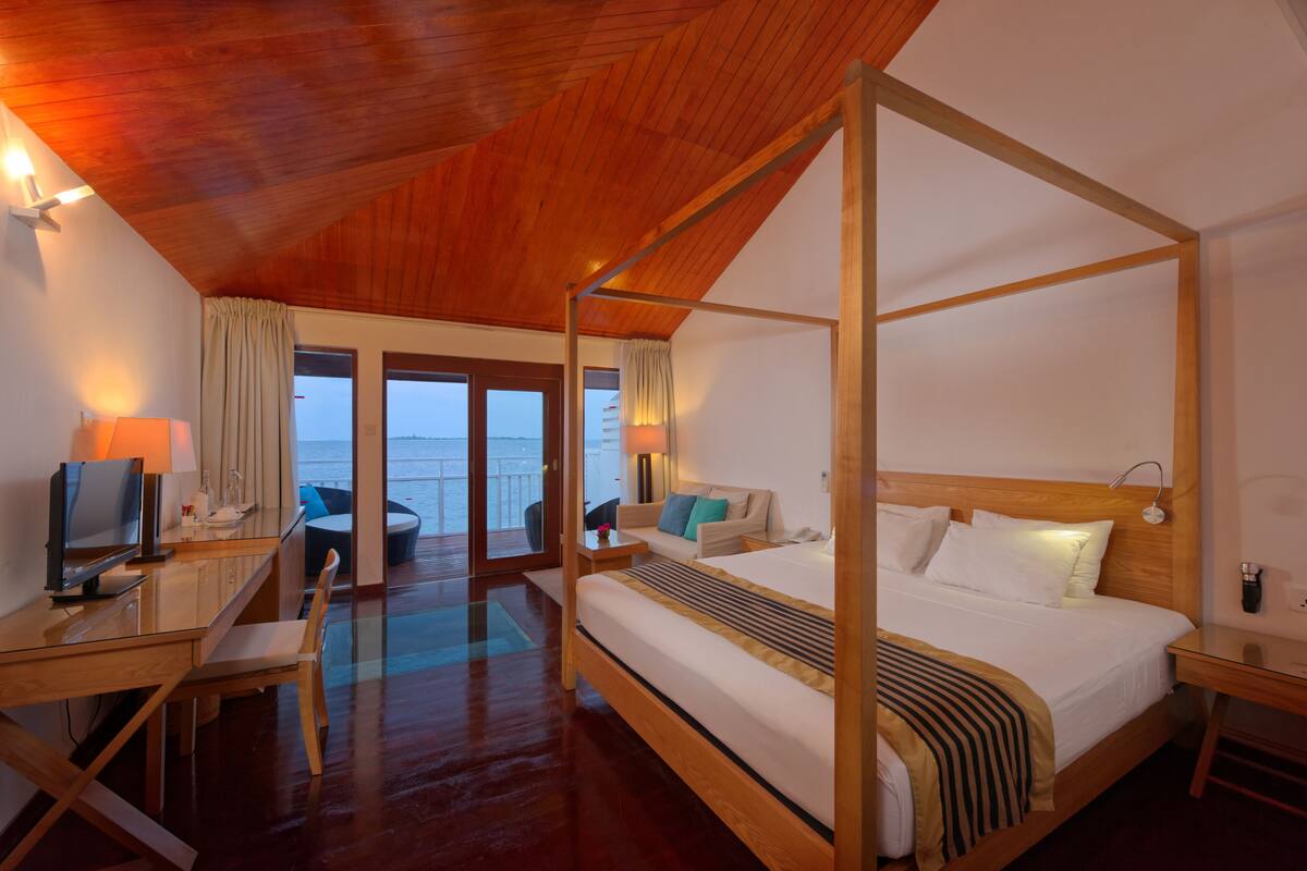 Una cama con dosel de madera, un escritorio con una silla, un televisor y vistas al mar a través de puertas de cristal.