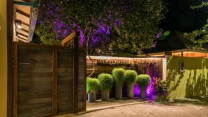 Una entrada ben il·luminada a una propietat anomenada "Holiday Garden Retreat", amb una porta de fusta, plantes en testos i un arbre amb il·luminació morada.