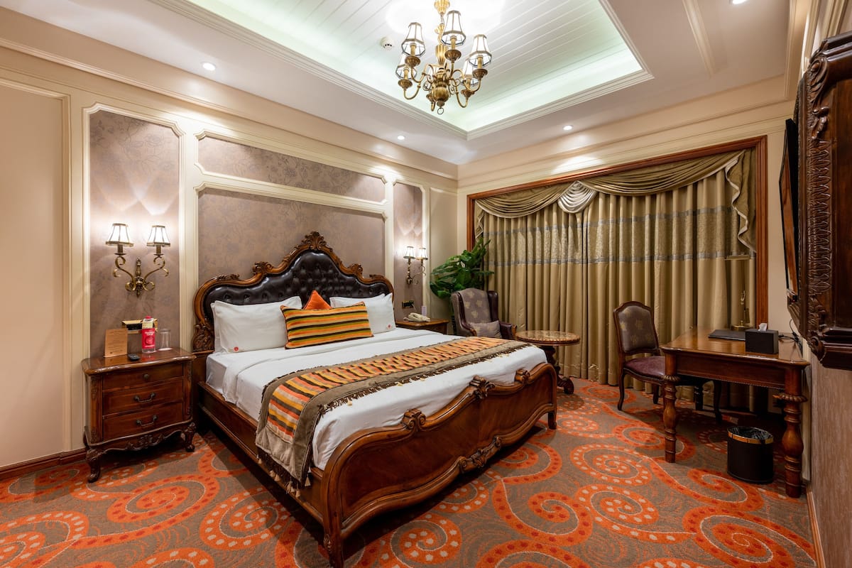 A large bed with ornate headboard, a nightstand, a desk with a chair, and a potted plant.