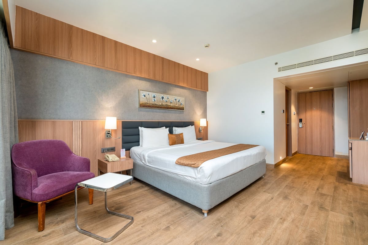 A modern hotel room with a bed, a purple armchair, a small table, and a painting on the wall.