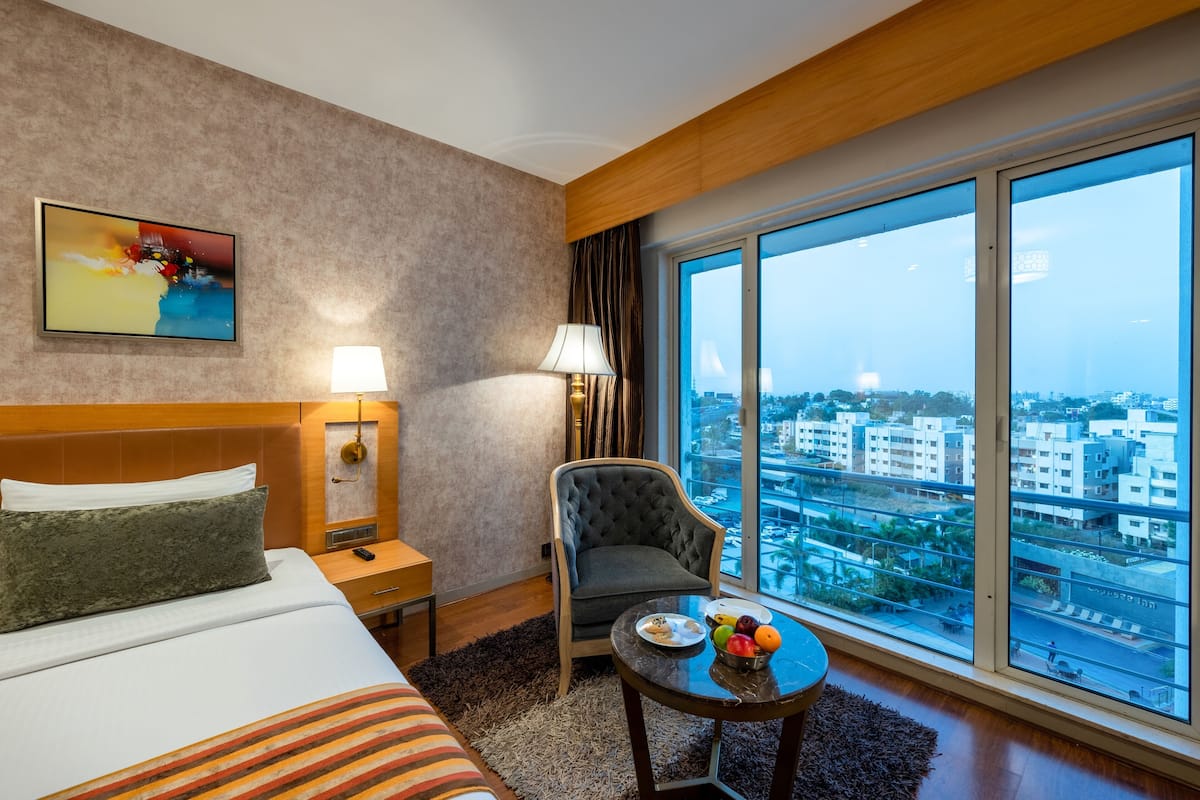 A hotel room with a bed, a chair, a table with fruit, a lamp, and a large window with a city view.