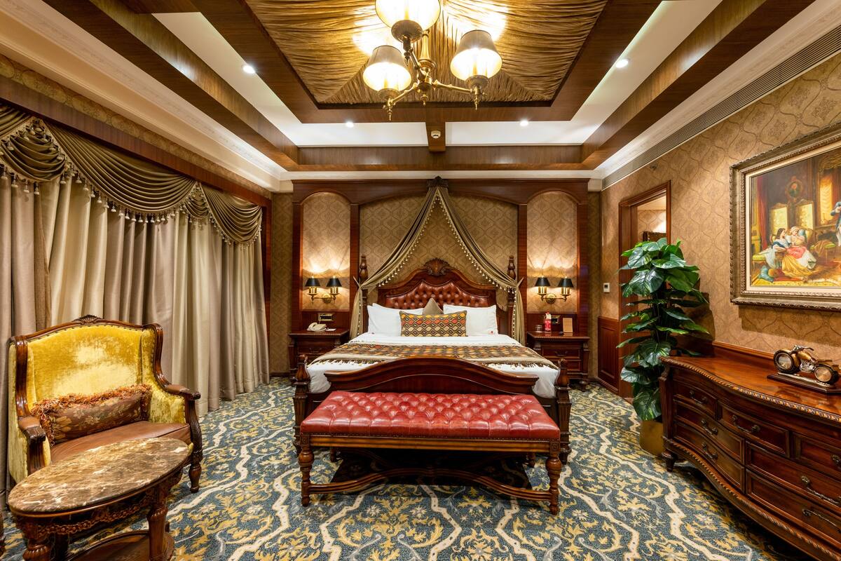 A luxurious bedroom with a large bed, ornate headboard, a sitting chair, a bench, a large potted plant, and a classic clock on a dresser.