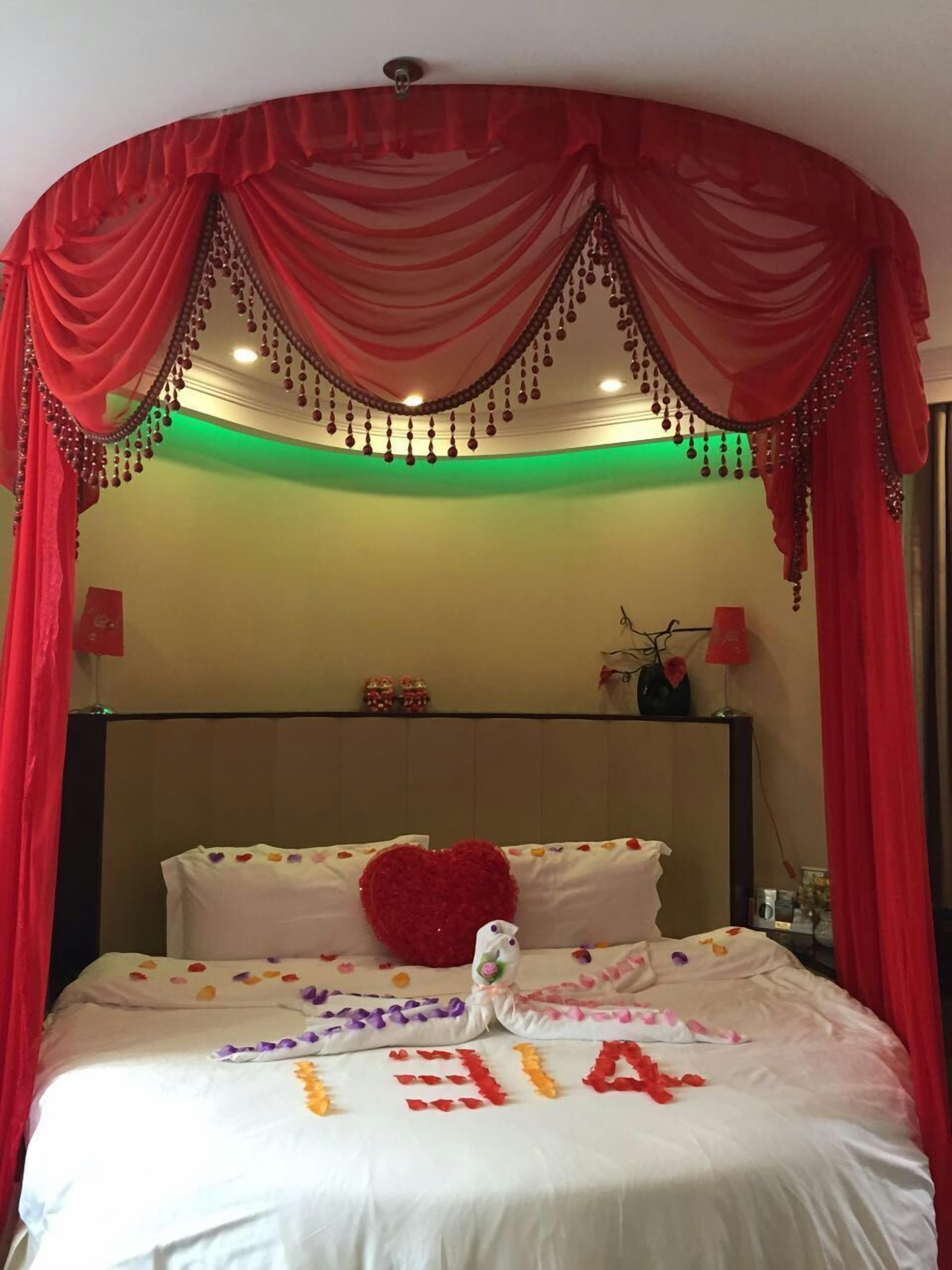 A hotel room with a bed adorned with a heart-shaped pillow, a floral arrangement, and a message spelled out with rose petals.