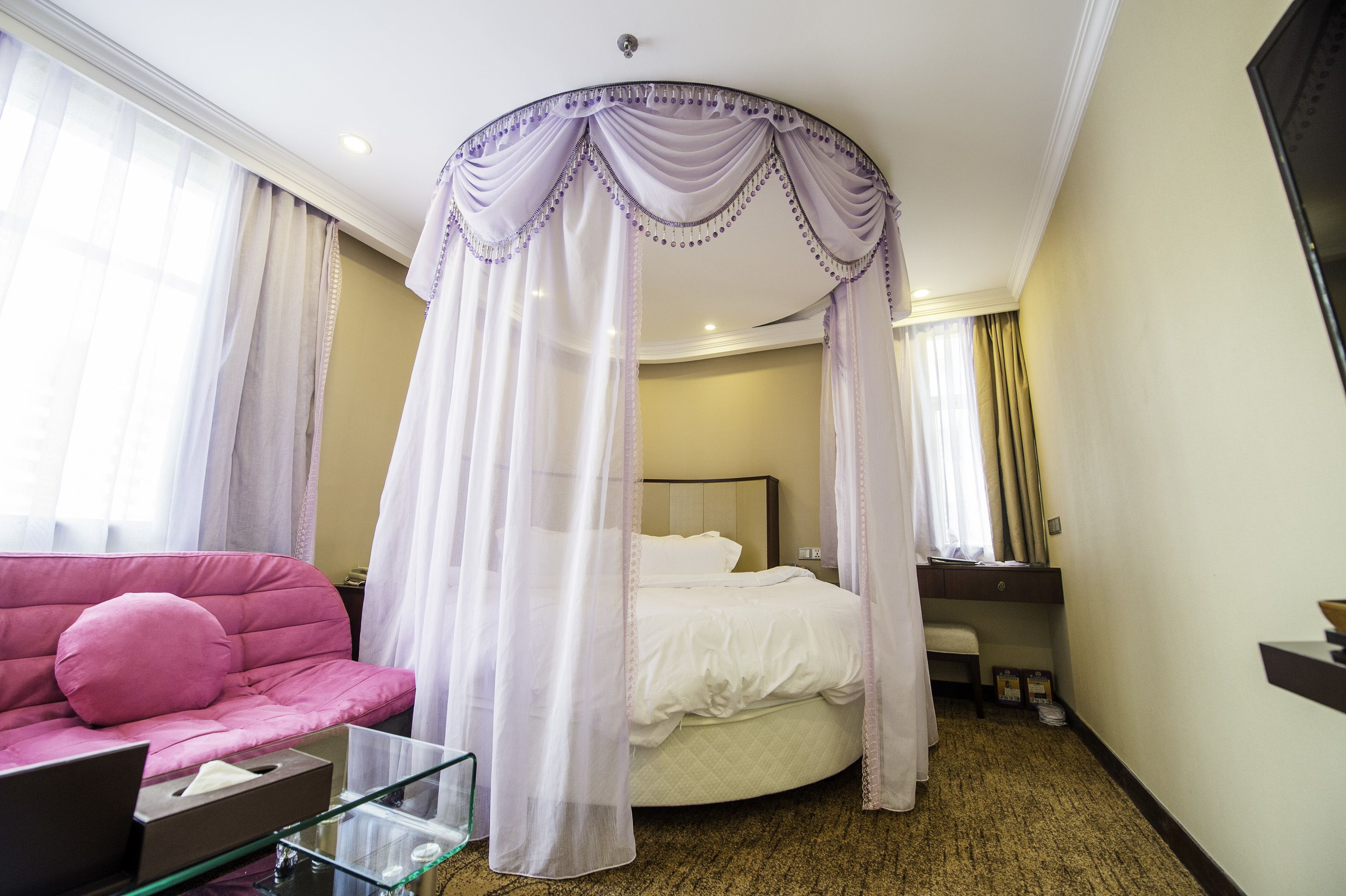 A hotel room with a bed, a pink sofa, a glass coffee table, and a TV on the wall.