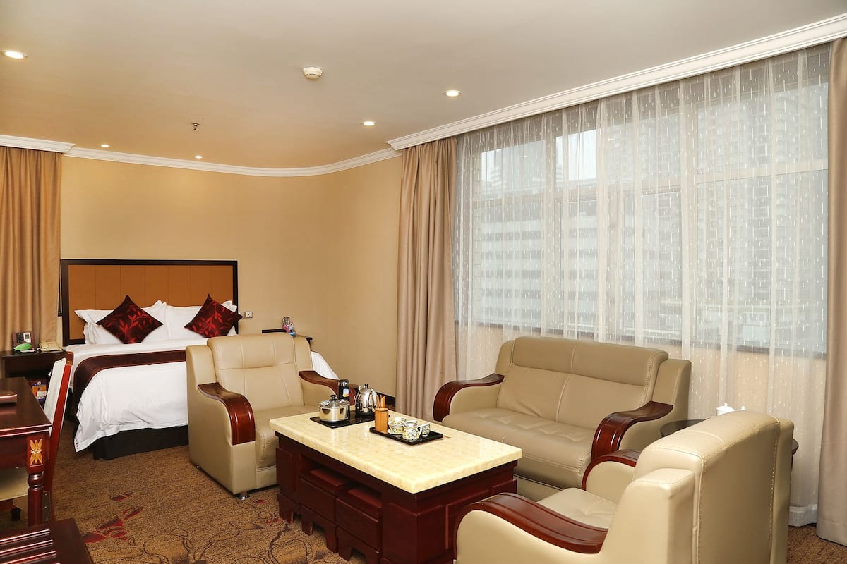 Een hotelkamer met een bed, twee fauteuils, een bijzettafeltje en een groot raam met dunne gordijnen.