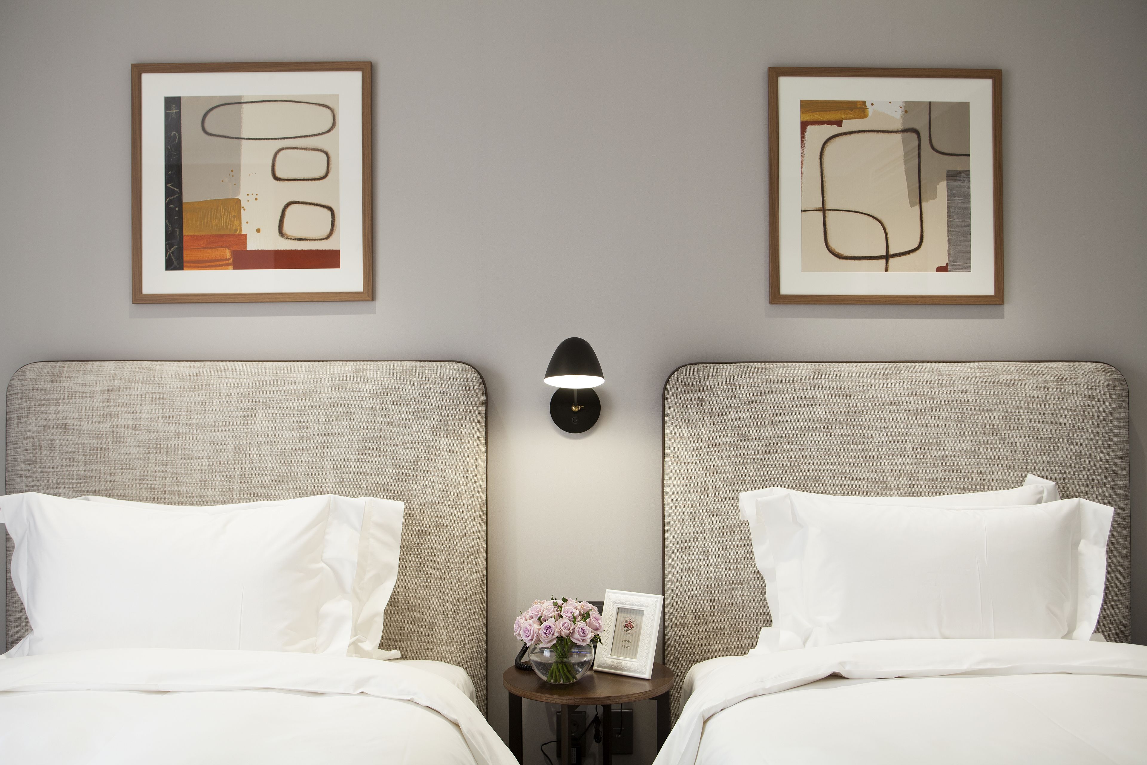 Une chambre d’hôtel avec deux lits, une table de chevet avec une lampe et deux œuvres d’art abstraites encadrées accrochées au mur.