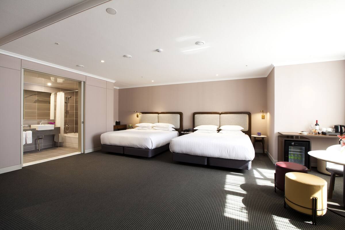 A hotel room with two beds, a desk, a chair, a small table, a refrigerator, and a bathroom with a shower and sink.