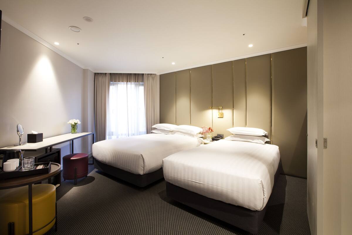A hotel room with two beds, a desk with a chair, a small stool, and a mirror.
