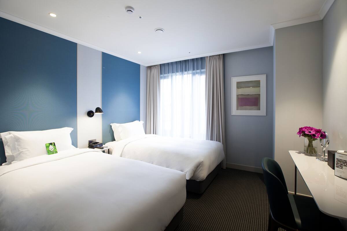 A hotel room with two beds, a desk, a chair, a window with curtains, a wall-mounted lamp, and a framed picture.