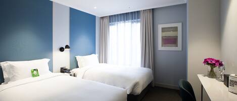 Habitación de hotel con dos camas, un escritorio, una silla, una ventana con cortinas, una lámpara de pared y un cuadro enmarco.
