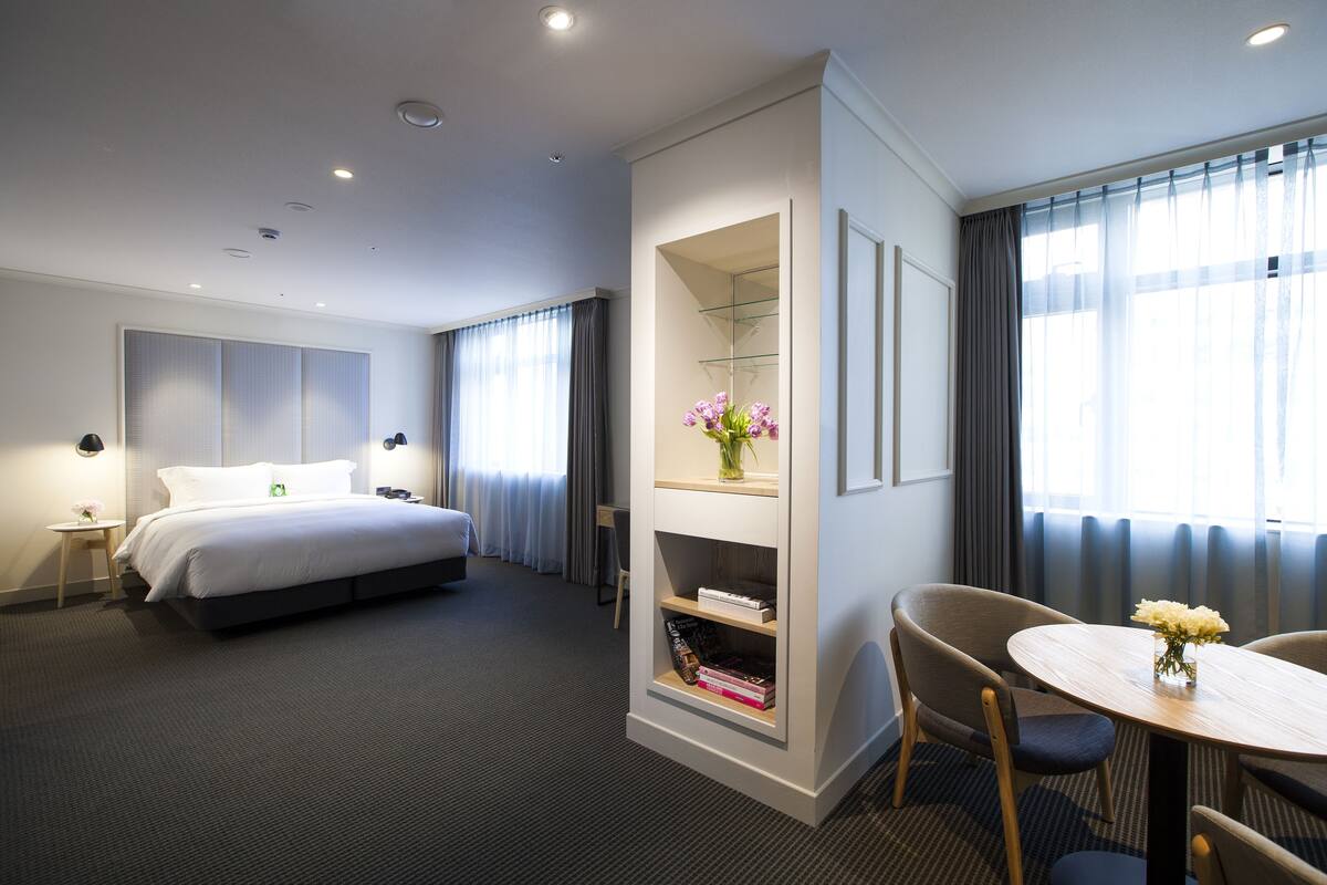 A hotel room with a bed, a desk, a chair, a small table with a vase, and a bookshelf.