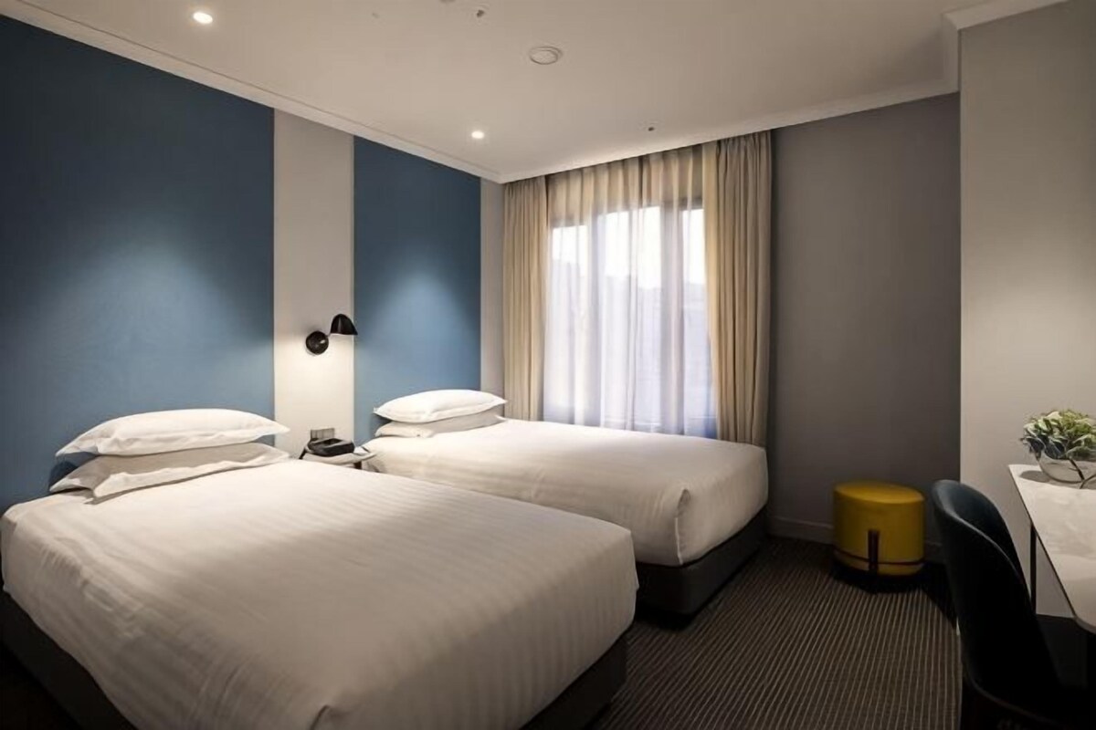 A hotel room with two beds, a desk, a chair, a small table, and a window with curtains.