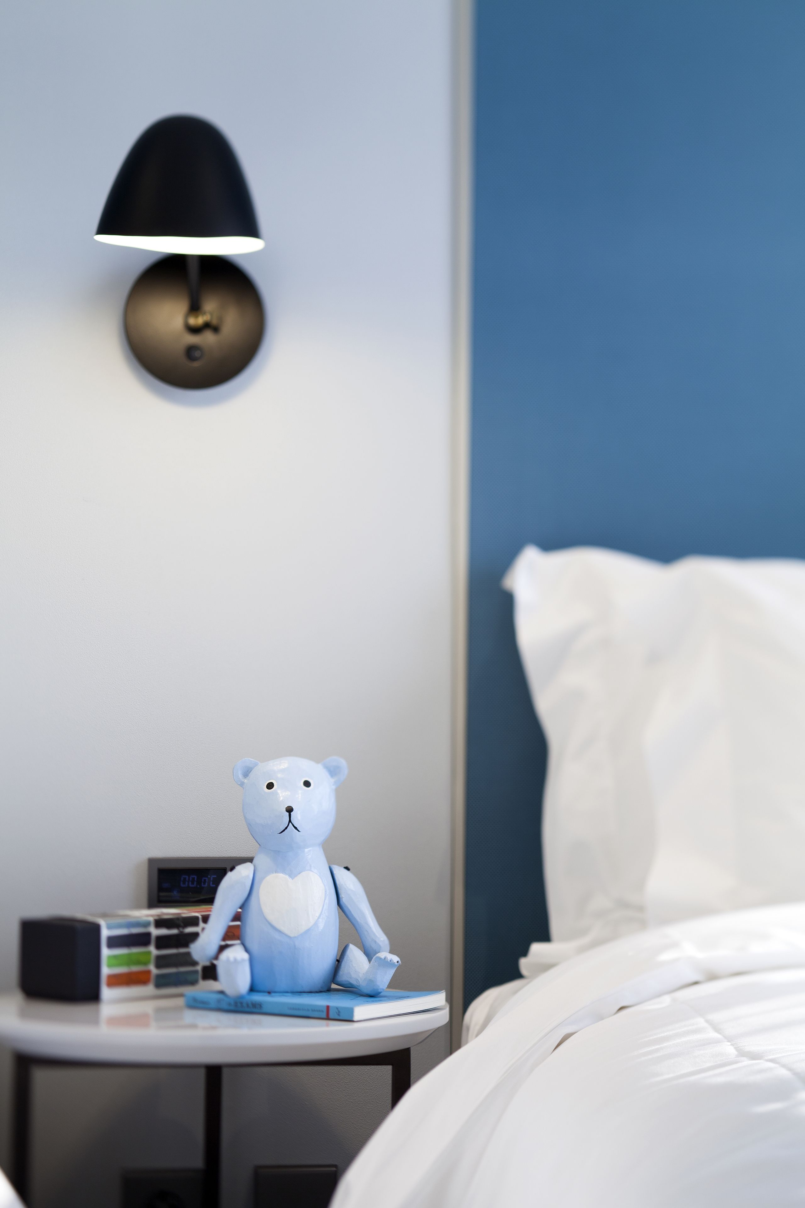 A bedroom with a bed, a bedside table with a lamp, a teddy bear, and a digital clock.