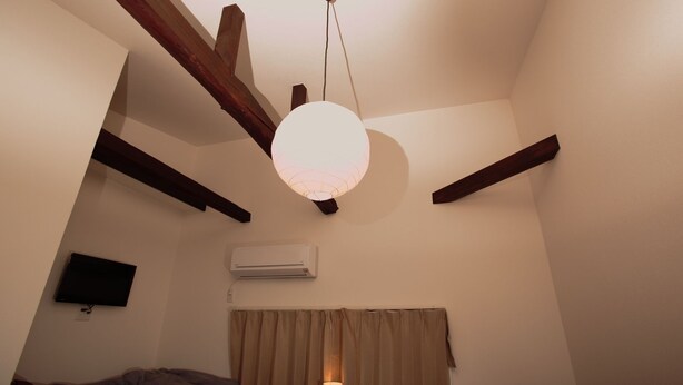 A room with a sloped ceiling, exposed wooden beams, a hanging light fixture, a flat-screen TV, an air conditioning unit, and a window with curtains. - Place Iine (Tokyo)