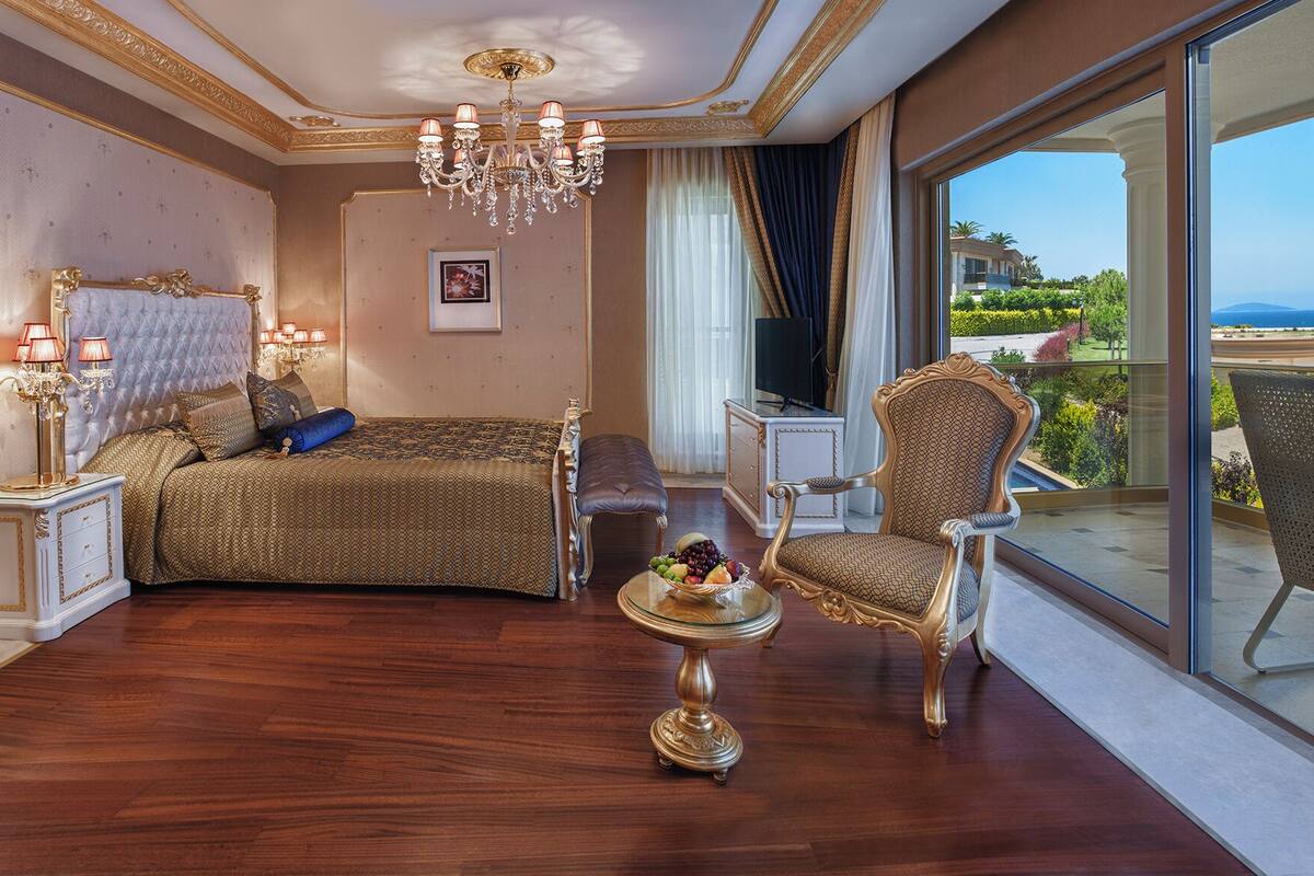 A luxurious bedroom with a large bed, a sitting area with a chair and a table, and a balcony with a view.