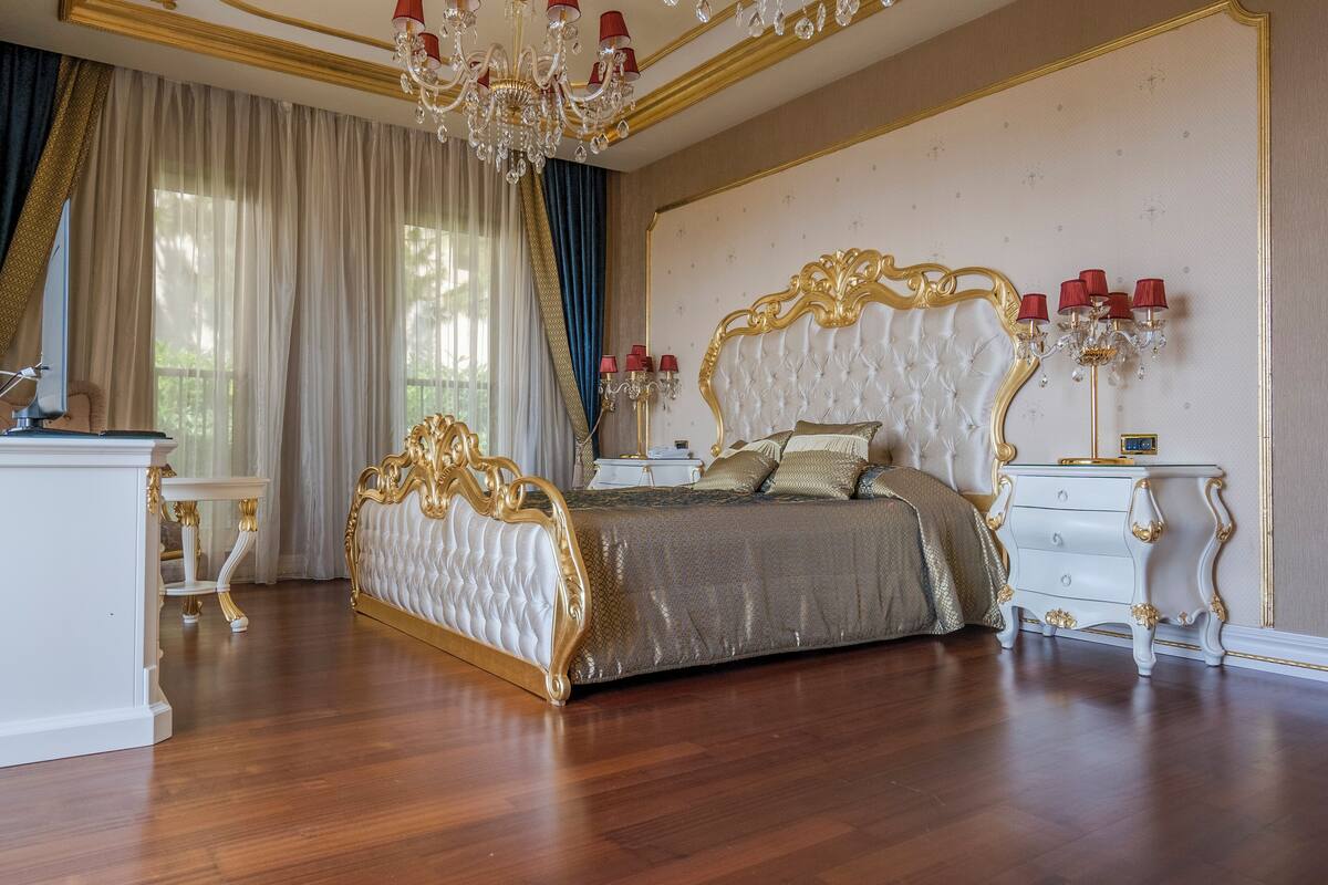 A luxurious bedroom with a large bed, ornate gold detailing, a chandelier, and a nightstand with red lamps.