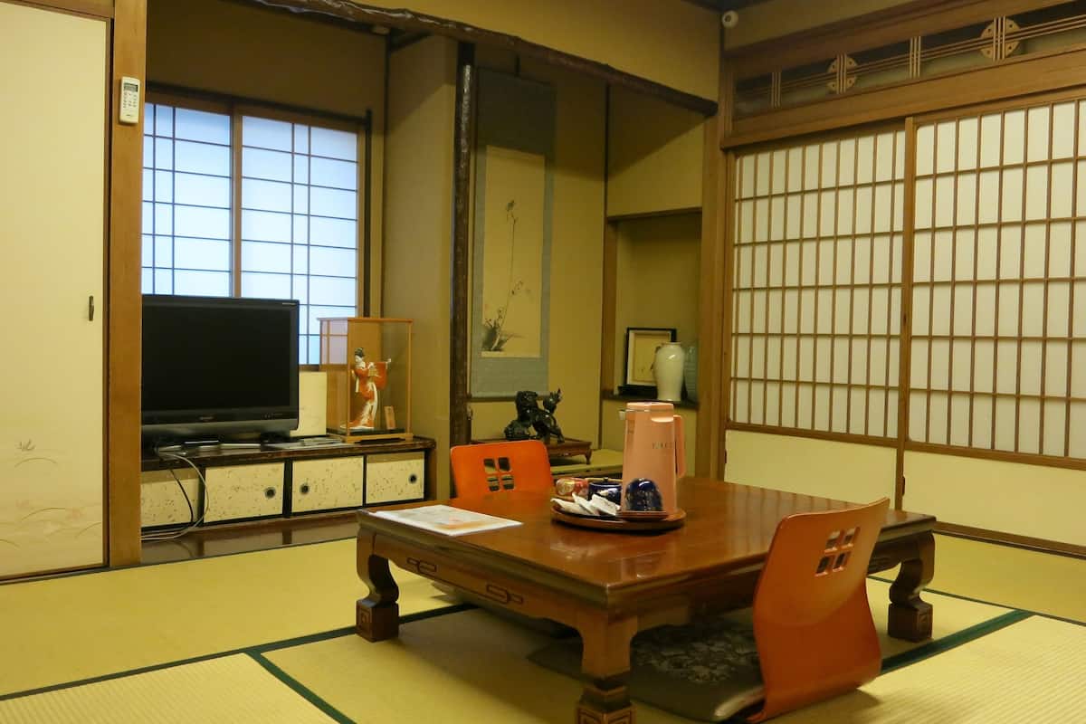 Una habitación japonesa tradicional con suelo de tatami, una mesa de madera baja, sillas naranjas, un televisor y puertas correderas con paneles shoji.