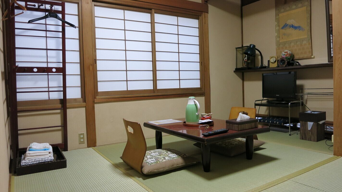 Una habitación japonesa tradicional con suelo de tatami, una mesa baja con cojín, un televisor y una estantería con adornos.