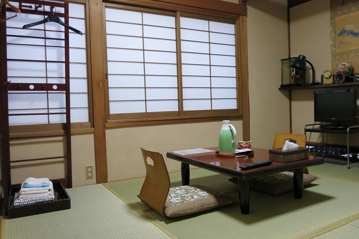 Una habitación japonesa tradicional con suelo de tatami, una mesa baja con cojín, un televisor y una estantería con adornos.