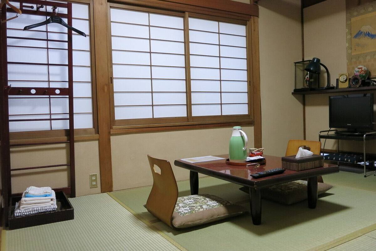 Una habitación japonesa tradicional con suelo de tatami, una mesa baja con cojín, un televisor y una estantería con adornos.