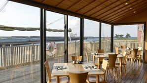 An outdoor dining area with wooden tables and chairs, large windows offering a view of the sea and a distant building. - Kasasa Ebisu (Minamisatsuma)
