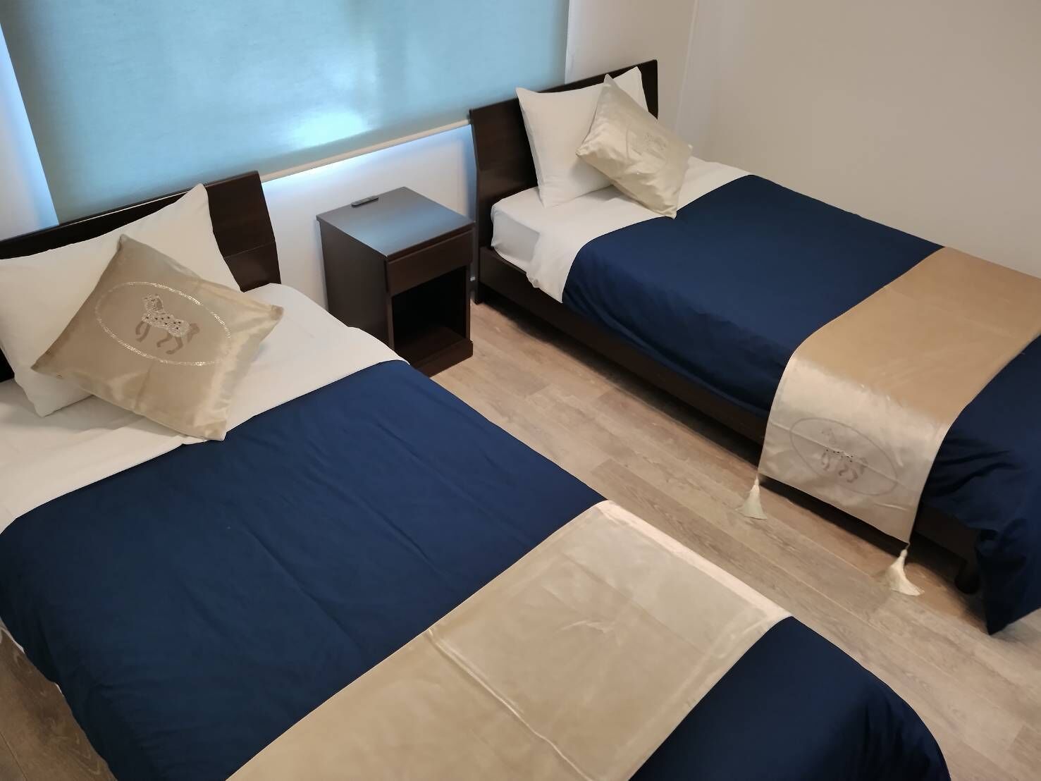 Ett hotellrum med två sängar, var och en med ett blått överkast och en beige kudde. Mellan sängarna står ett nattduksbord, och det finns ett fönster med utsikt över himlen.
