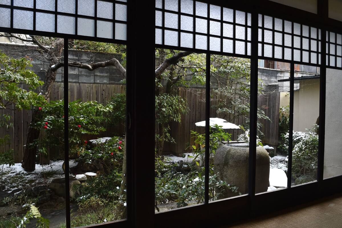 Une pièce de style japonais traditionnel, avec un sol recouvert de tatamis, un paravent en papier de riz (shoji) et une vue sur un jardin agrémenté d’une clôture en bois, de rochers et de plantes.