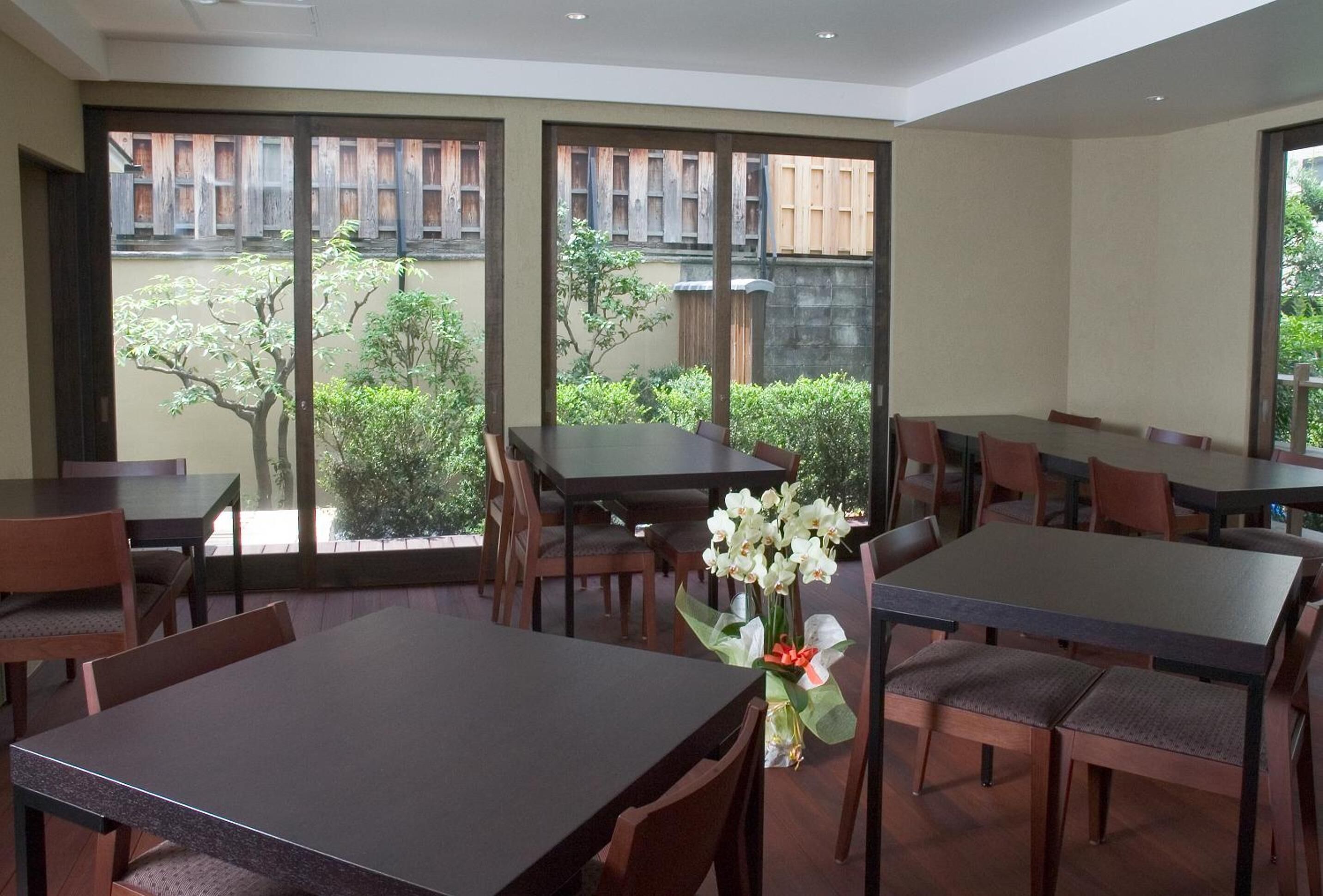 A dining area with multiple tables and chairs, large windows with a view of greenery and a wooden fence.