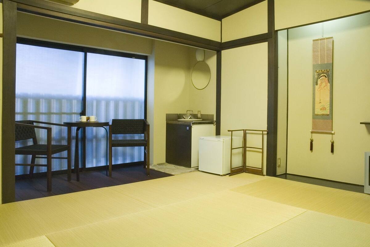 Une pièce de style japonais traditionnel, avec un sol recouvert de tatamis, une petite table basse entourée de chaises, un petit réfrigérateur et une porte coulissante donnant sur un balcon avec vue.