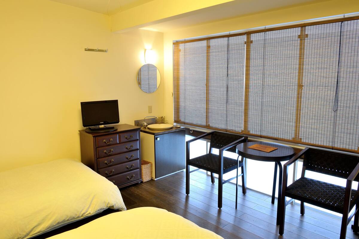 Une chambre avec un lit, une commode, une télévision, un petit lavabo et une fenêtre avec des stores.