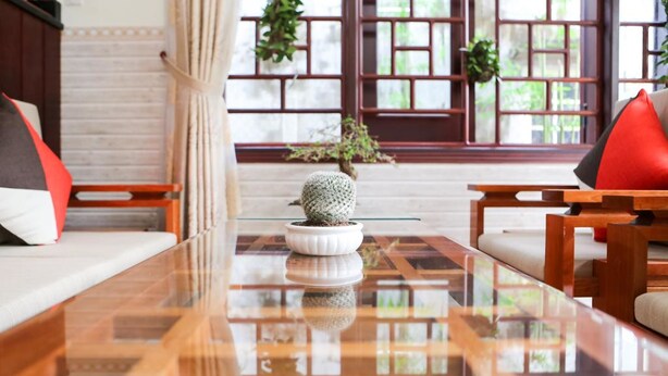 A modern living room with a glass coffee table, wooden furniture, and a view through a traditional window with plants. - Viet Nhat Homestay (Da Nang)