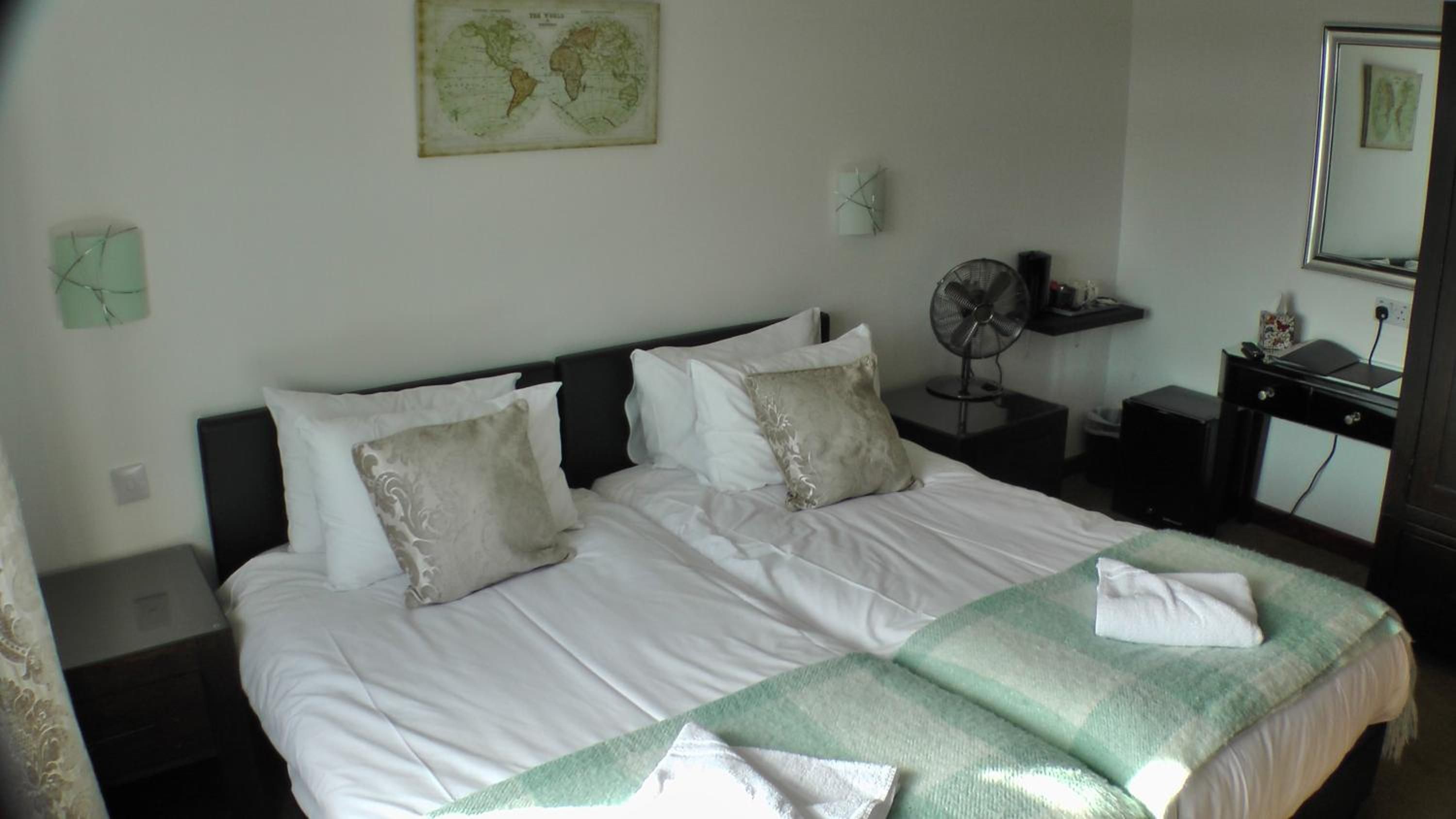 Una cama doble con sábanas blancas y dos almohadas, una mesita de noche con un ventilador, un estante con objetos y un mapa mundial en la pared.