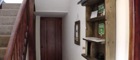 Un pasillo estrecho con una puerta de madera, un pequeño estante de madera con un letrero verde y una escalera con barandillas de madera.