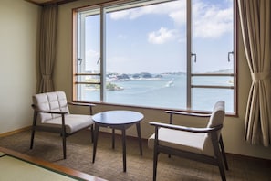 A room with a large window offering a view of the sea and distant land. There are two chairs and a small round table. - Toba Seaside Hotel (Toba)