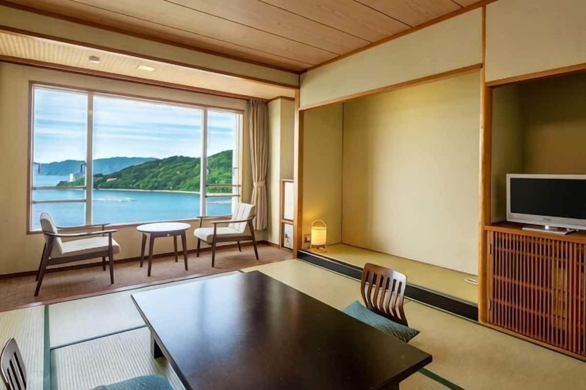 A hotel room with a large window, a dining table, chairs, a TV, and a traditional Japanese-style cabinet.
