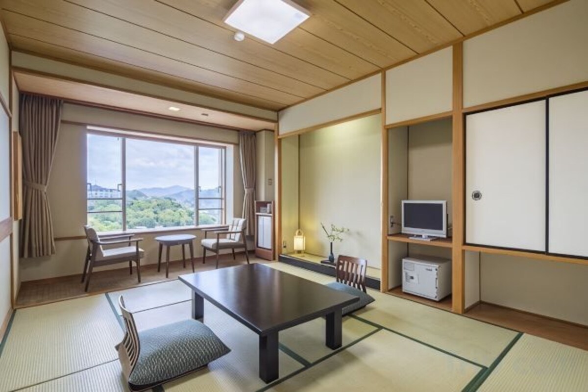 Una habitación japonesa tradicional con suelo de tatami, una mesa baja, sillas, televisor y vistas al exterior.