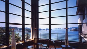A high-ceilinged room with large windows offering a view of a body of water and distant buildings. - Toba Seaside Hotel (Toba)