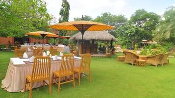 An outdoor dining area with tables and chairs, surrounded by lush greenery and a thatched-roof structure.