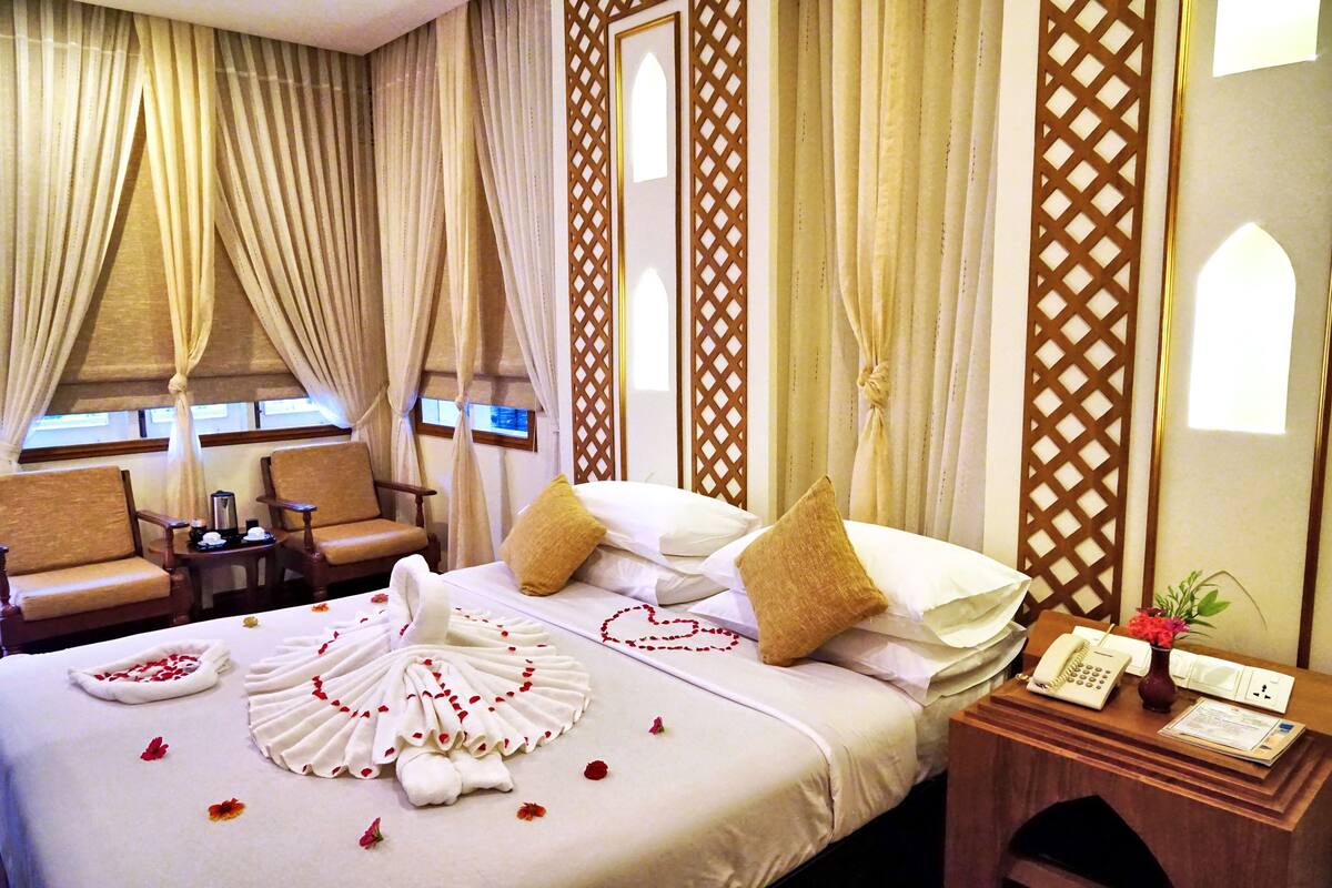 A hotel room with a neatly made bed, a wooden nightstand with a telephone and a vase of flowers, two chairs, and a window with curtains.