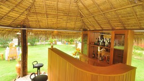 A bar area with a wooden counter and a glass display cabinet, surrounded by a thatched roof and outdoor seating. - KMA Bagan Village Hotel (Nyaung-U)