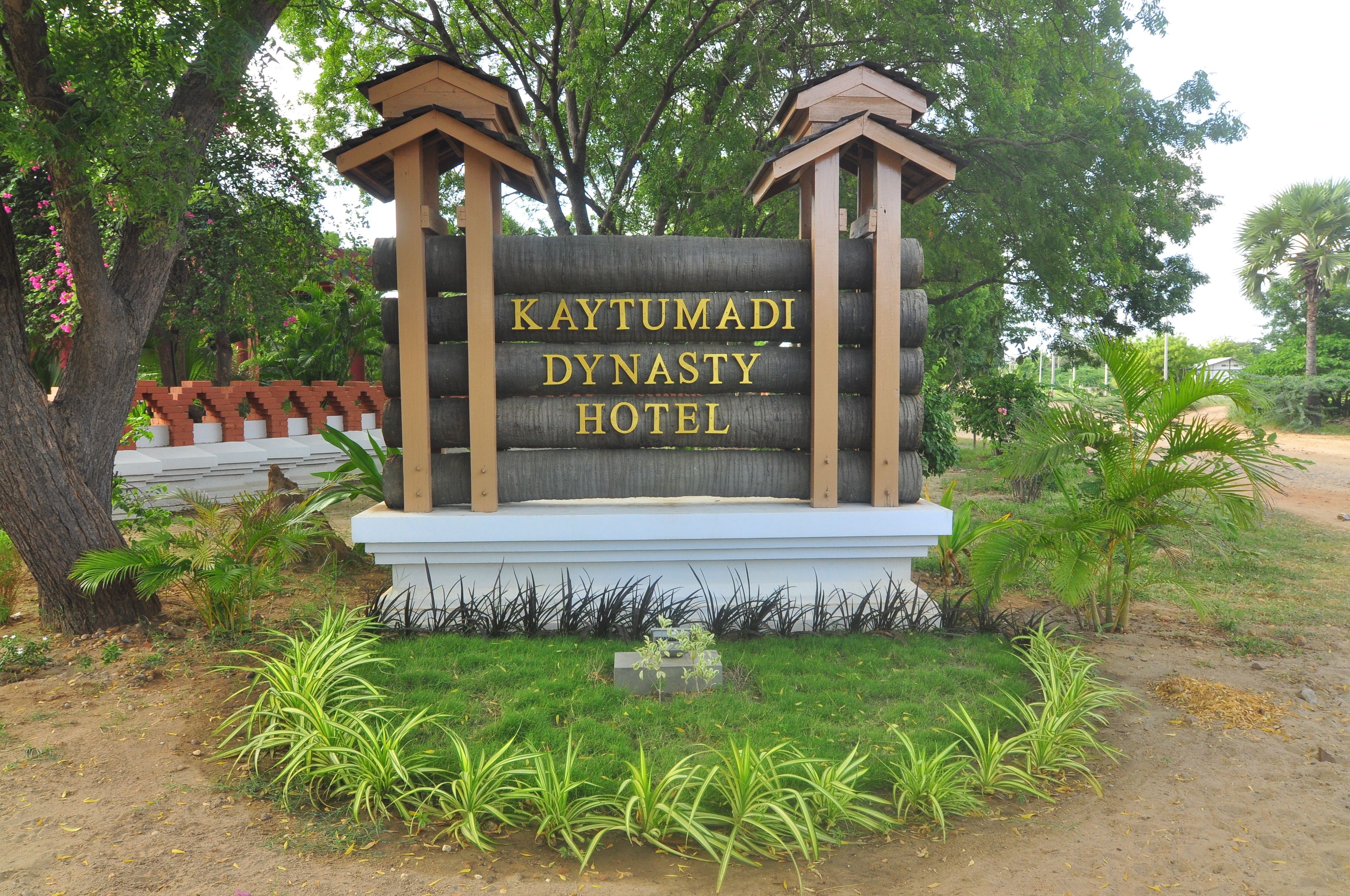 A sign for the Kaytumadi Dynasty Hotel with a wooden structure and a white base, surrounded by greenery and trees.
