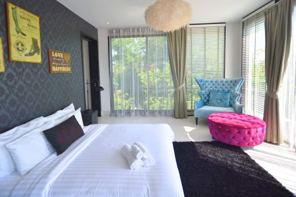 A bedroom with a large bed, a blue armchair, a pink ottoman, and a view of greenery through the window.