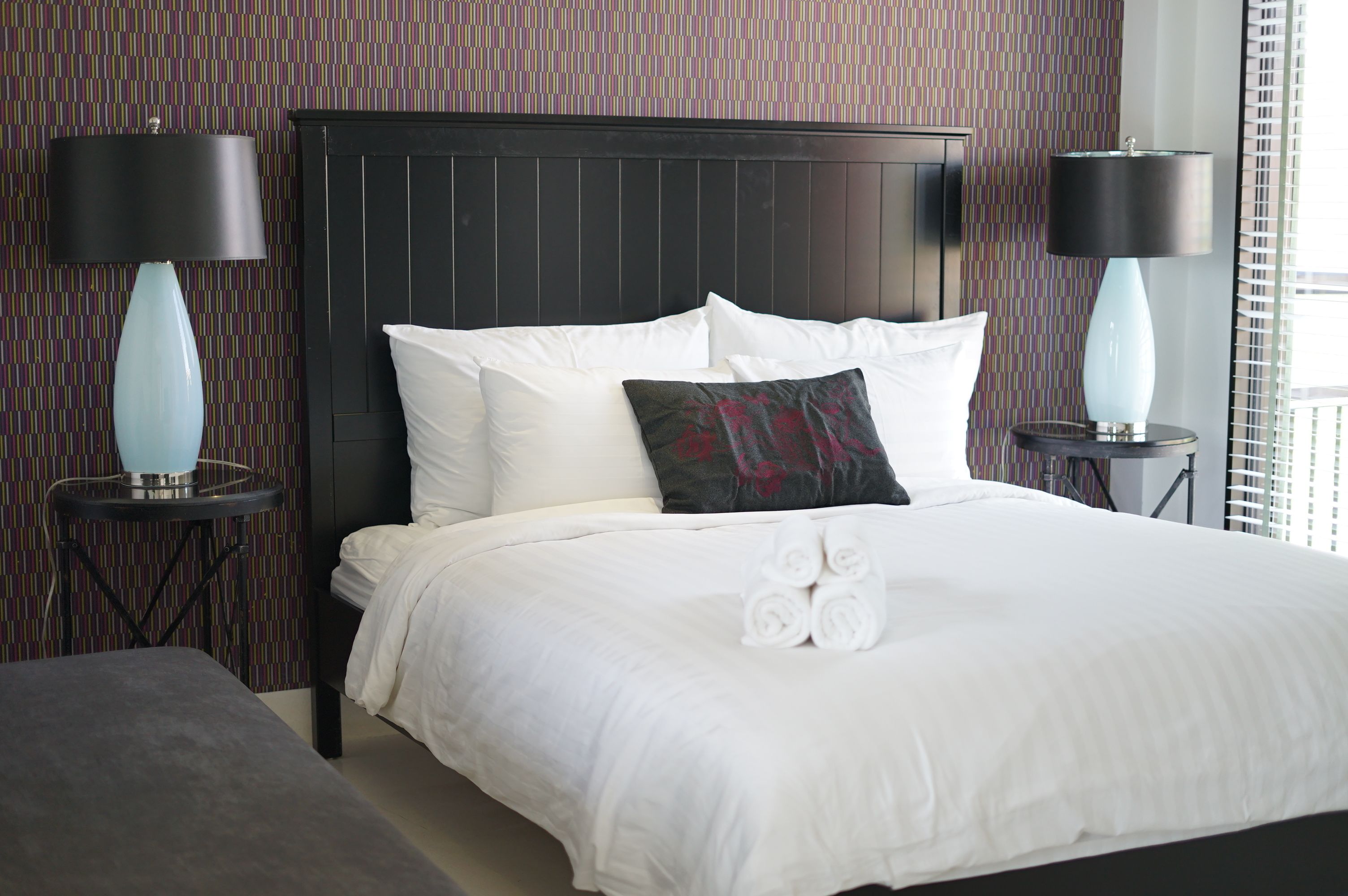 A neatly made bed with white linens and a decorative pillow, flanked by two black lamps on side tables, in a room with patterned wallpaper.