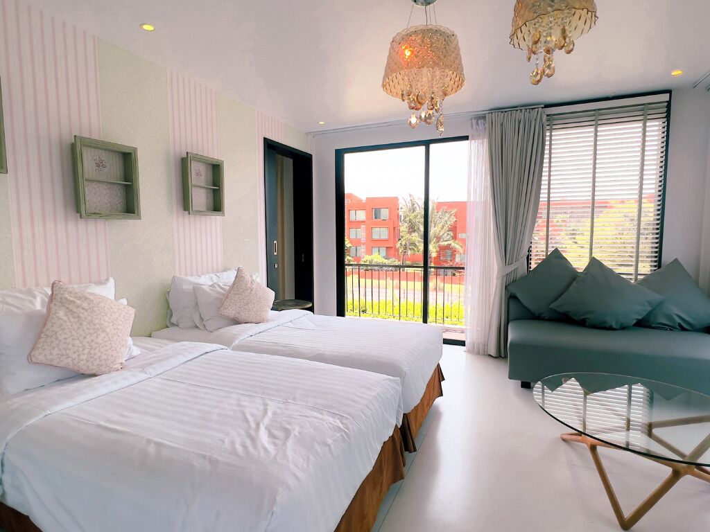 A hotel room with two beds, a sofa, a glass coffee table, and a large window with a view of buildings and greenery.