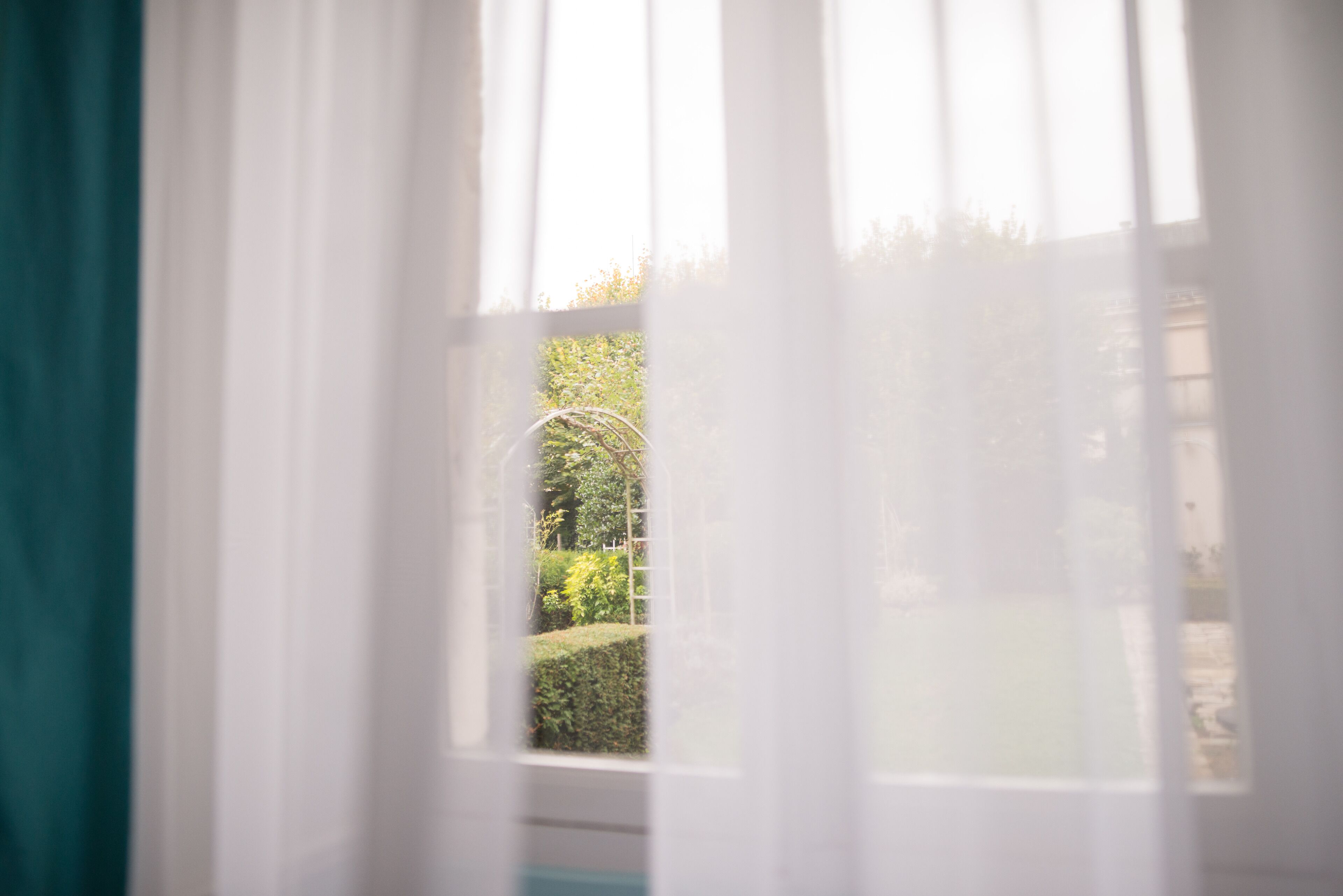 A window with sheer white curtains partially drawn, revealing a view of a garden with a hedge and a building in the background.